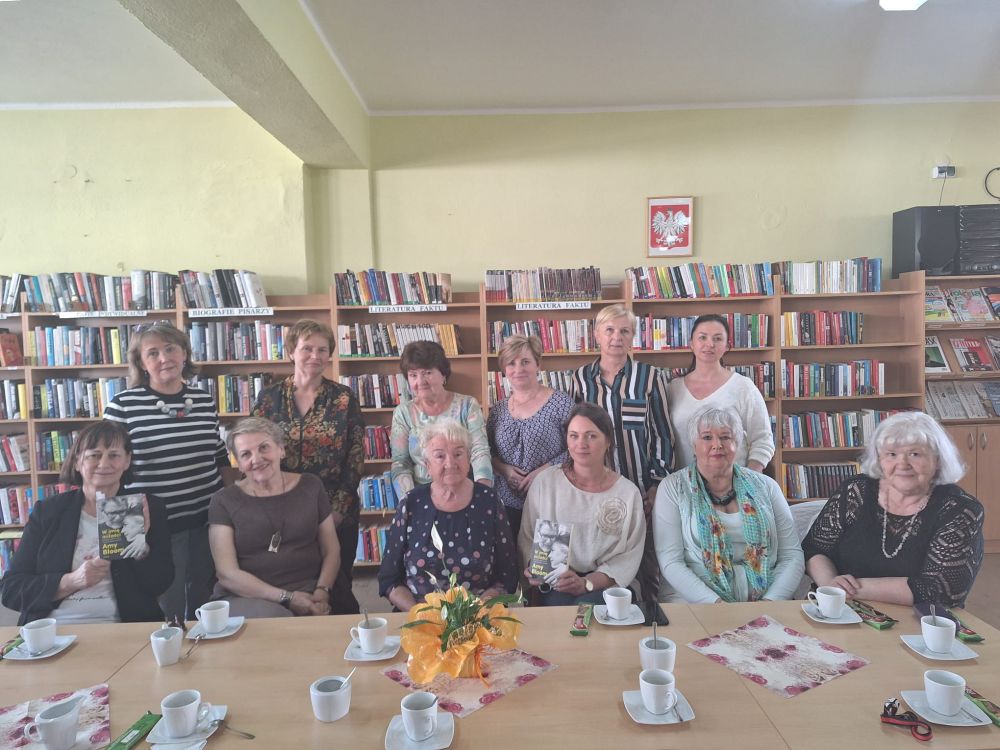 Uczestniczki spotkania DKK wraz z omawianą książką na tle regałów bibliotecznych.