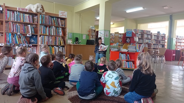 Dzieci siedząc na dywanie słuchają czytanej przez bibliotekarkę książki.