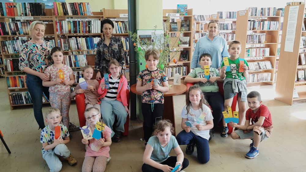 Grupa dzieci wraz z wykonanymi przez siebie pracami pozuje na tle regałów bibliotecznych.